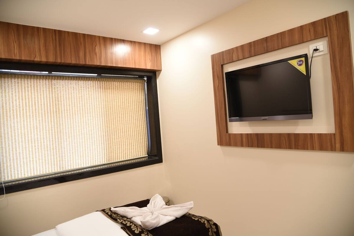 A hotel room with a large window, a flat-screen TV mounted on the wall, and a neatly made bed with a white blanket.