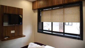Una habitación con una televisión instalada en una pared de paneles de madera, una ventana con persiana y una mesita de noche con un almohadón.