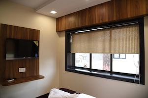 Una habitación con una televisión instalada en una pared de paneles de madera, una ventana con persiana y una mesita de noche con un almohadón.