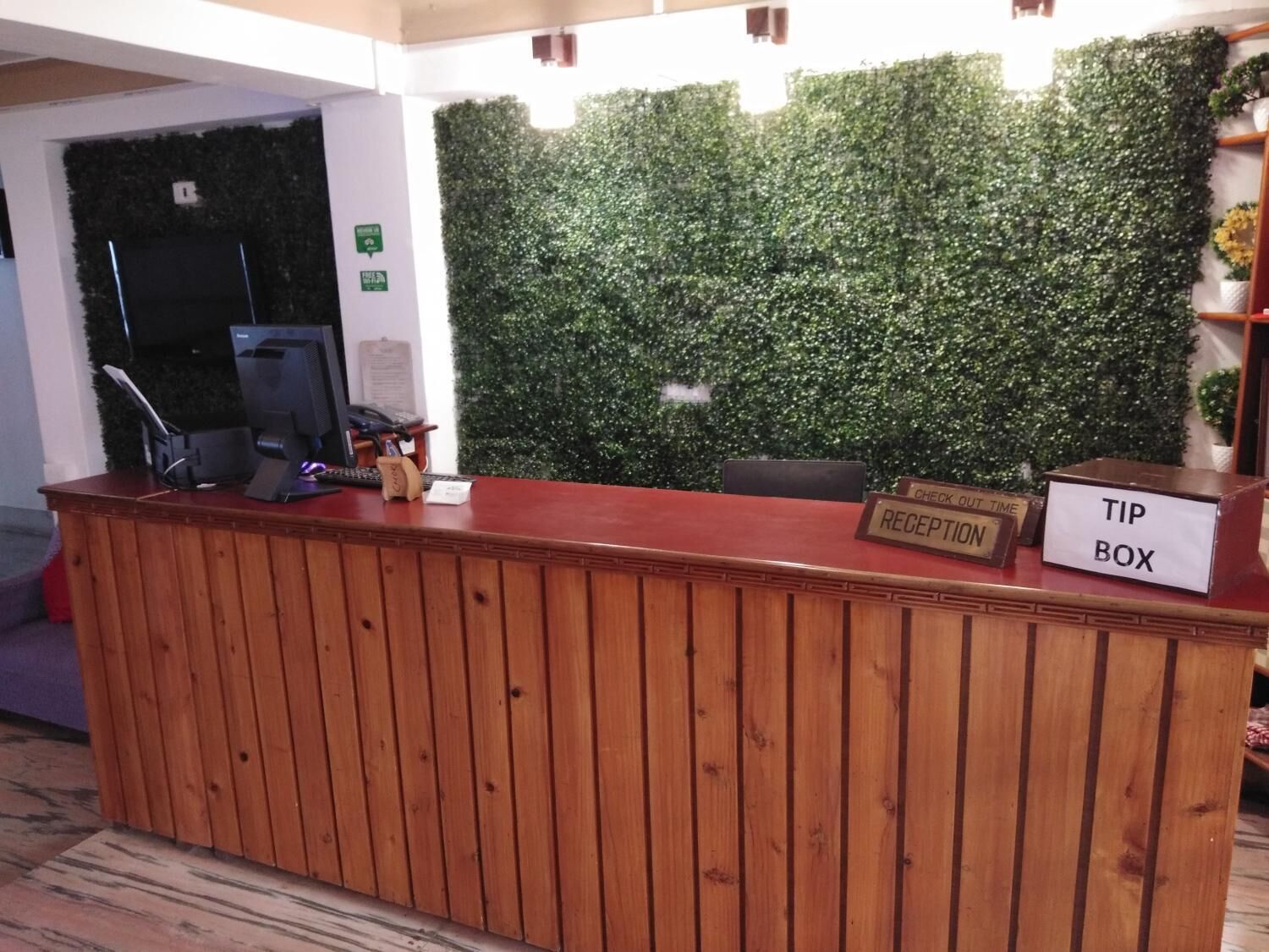 A reception desk with a wooden finish, a green wall with foliage, a computer, and a check-out time sign.