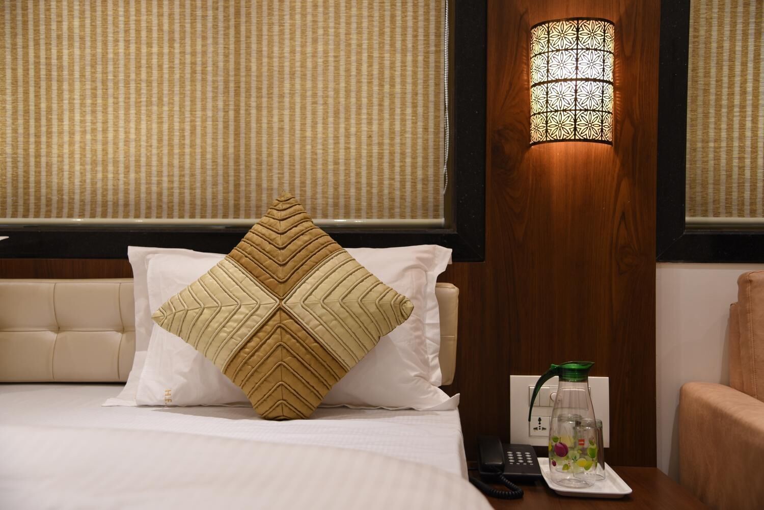 A hotel room with a bed, a nightstand with a phone and a glass pitcher, and a wall-mounted lamp.