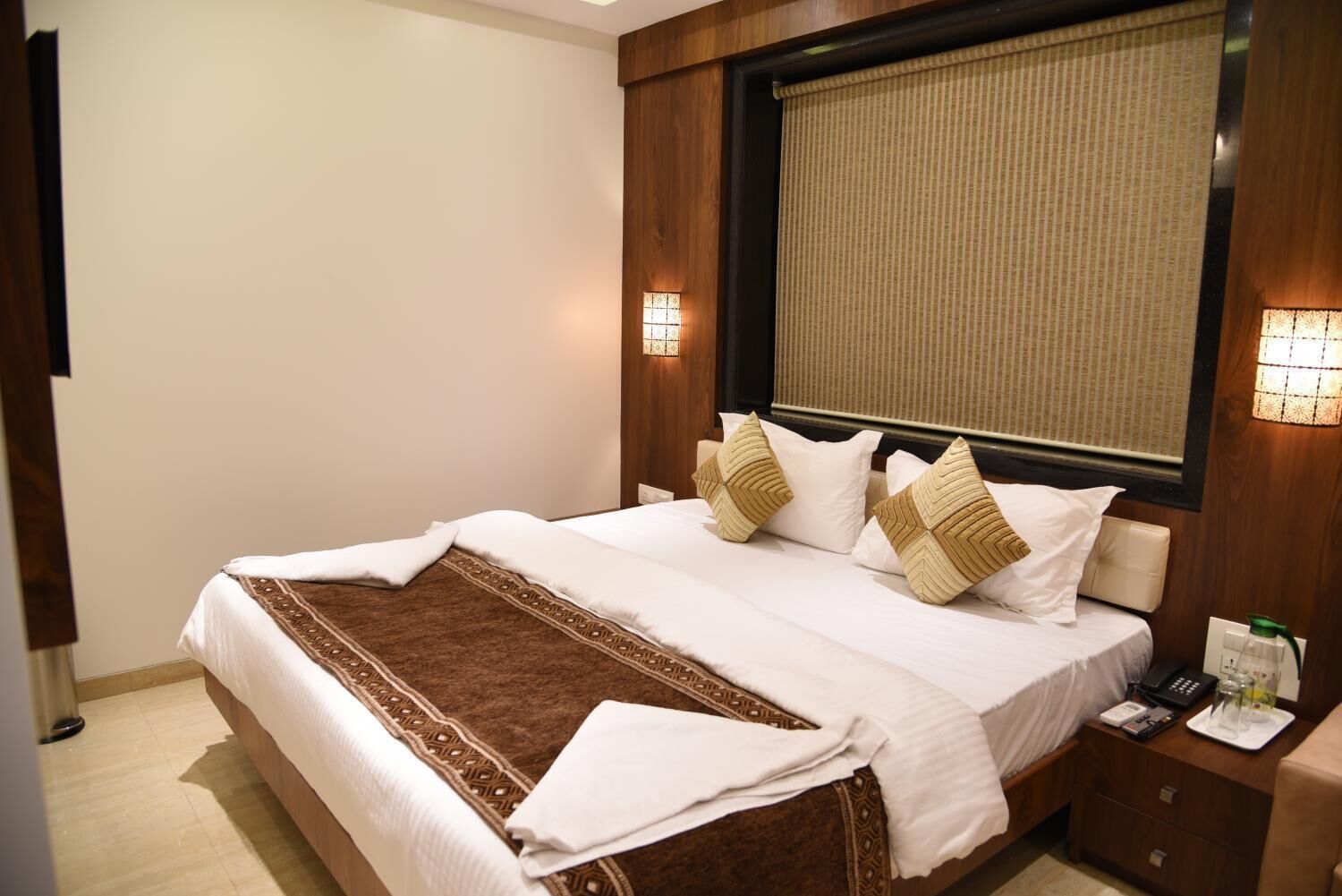 Cama bem arrumada com manta estampada, duas luminárias de cabeceira e mesa de cabeceira com telefone e copo.