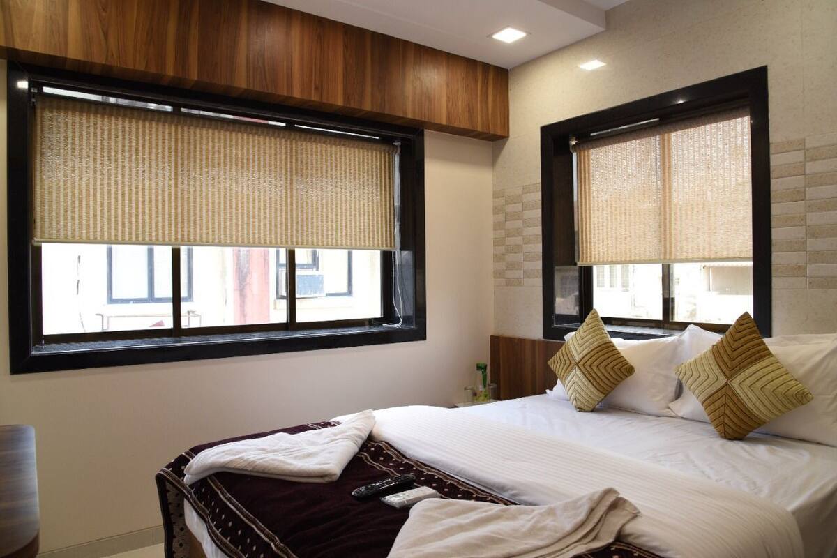 A hotel room with a bed, two windows with blinds, a wooden headboard, and a nightstand with a lamp.