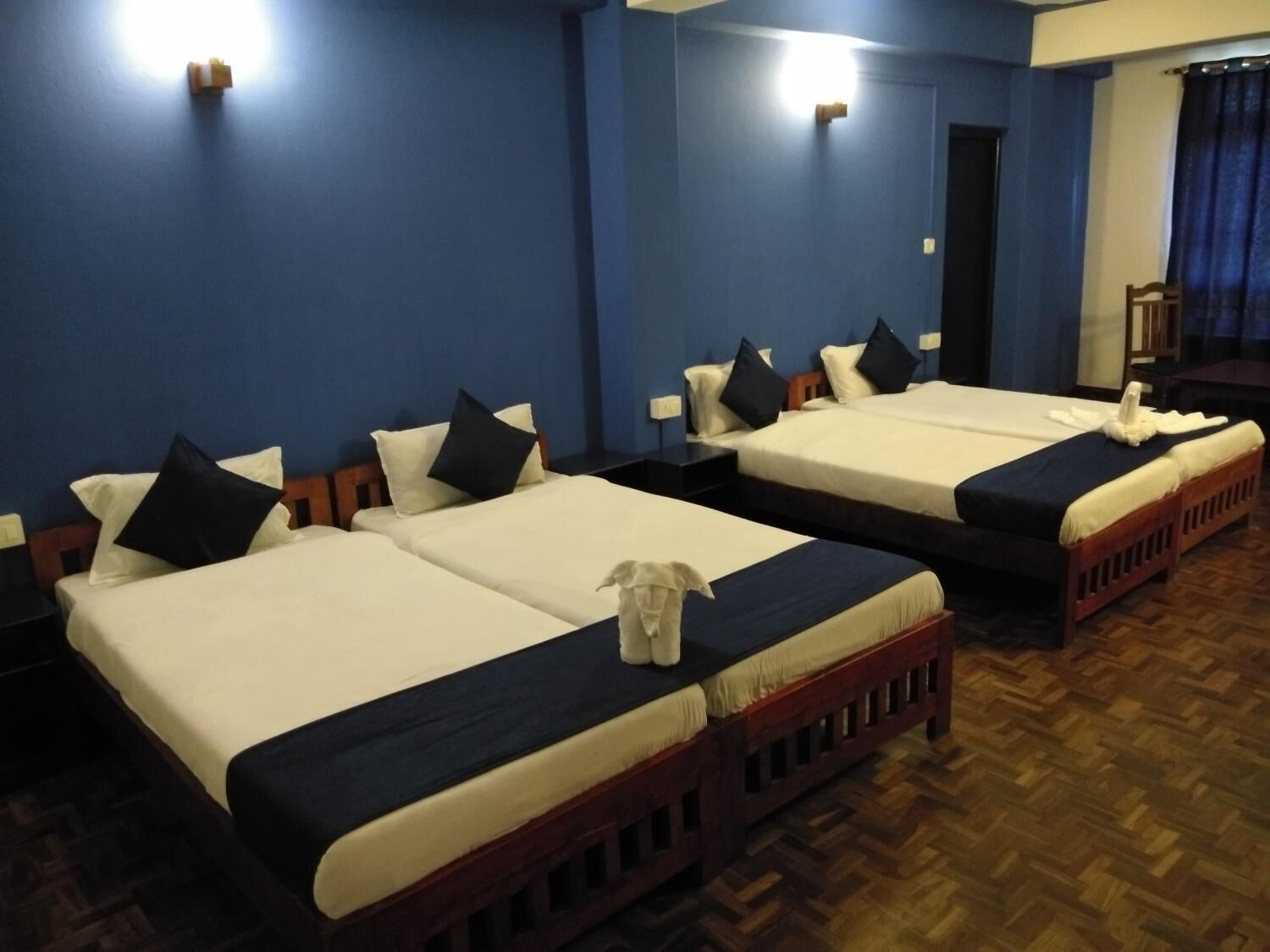 A hotel room with three single beds, each with a wooden frame and white bedding. There are two wall-mounted light fixtures on the blue walls.