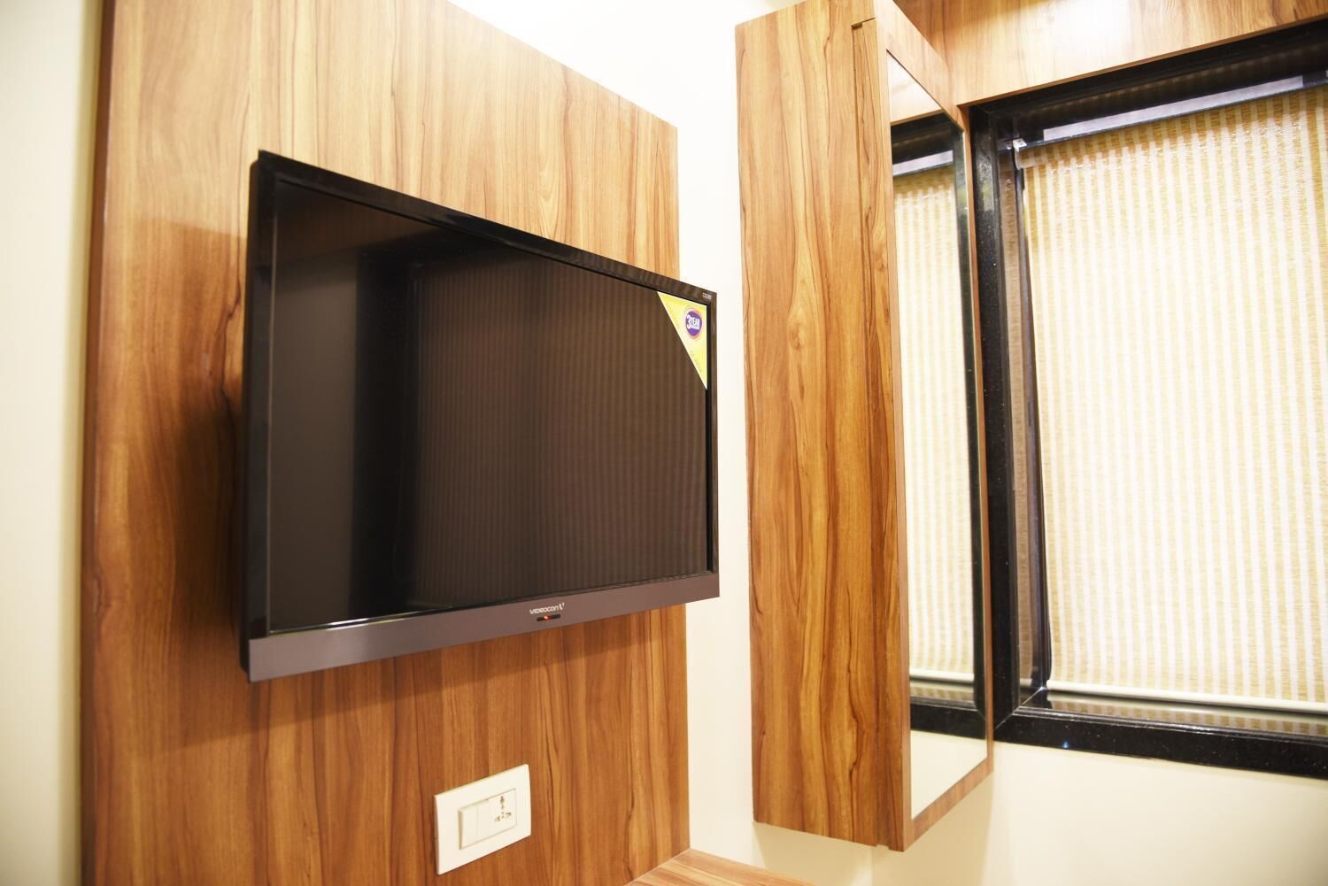 A flat-screen TV mounted on a wooden wall, a window with blinds, and a light switch.