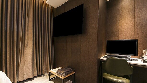 Una habitación de hotel con una cama, un escritorio con una computadora, una silla, una mesita con un cojín y un televisor montado en la pared.