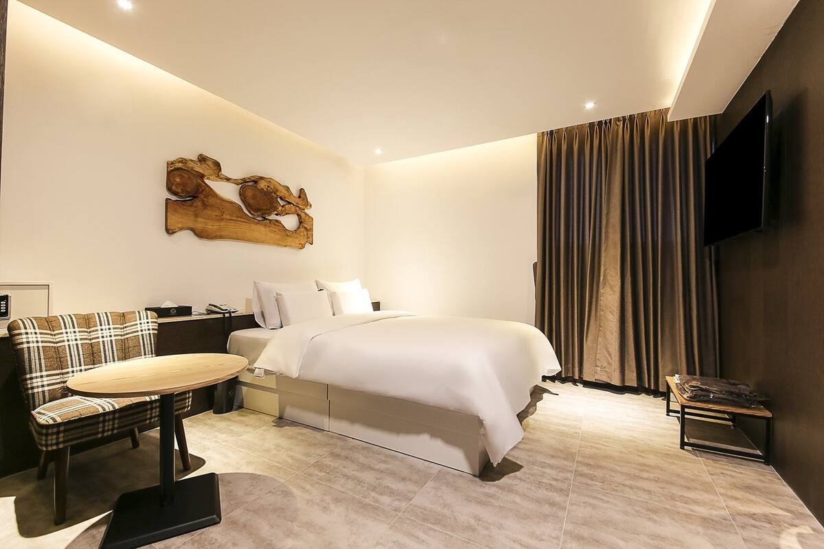 A modern hotel room with a large bed, a dining table, a sofa, a flat-screen TV, and a wooden art piece on the wall.