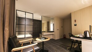 Habitación de hotel compacta con un pequeño comedor, un espejo, una silla de cuero negro, una mesa de madera y un cubo de basura negro.