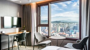 A hotel room with a large window offering a city view, a desk with a chair, a bed, and a TV mounted on the wall. - Hotel With Jeju (Jeju City)