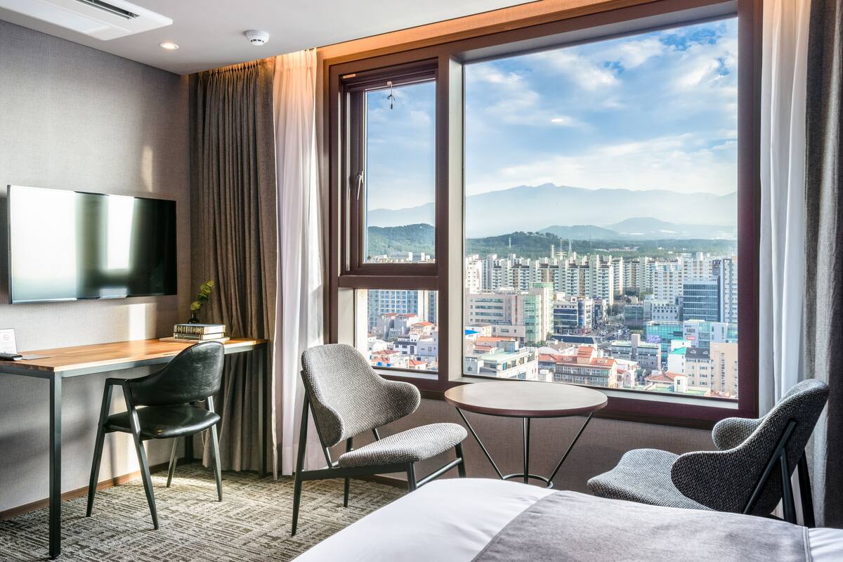 Une chambre d’hôtel dotée d’une grande fenêtre donnant sur la ville, d’un bureau avec une chaise, d’un lit et d’un téléviseur fixé au mur.