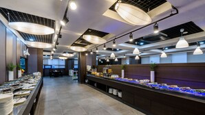 A buffet area with multiple food stations, overhead lighting, and a long counter with plates and utensils. - Hotel With Jeju (Jeju City)
