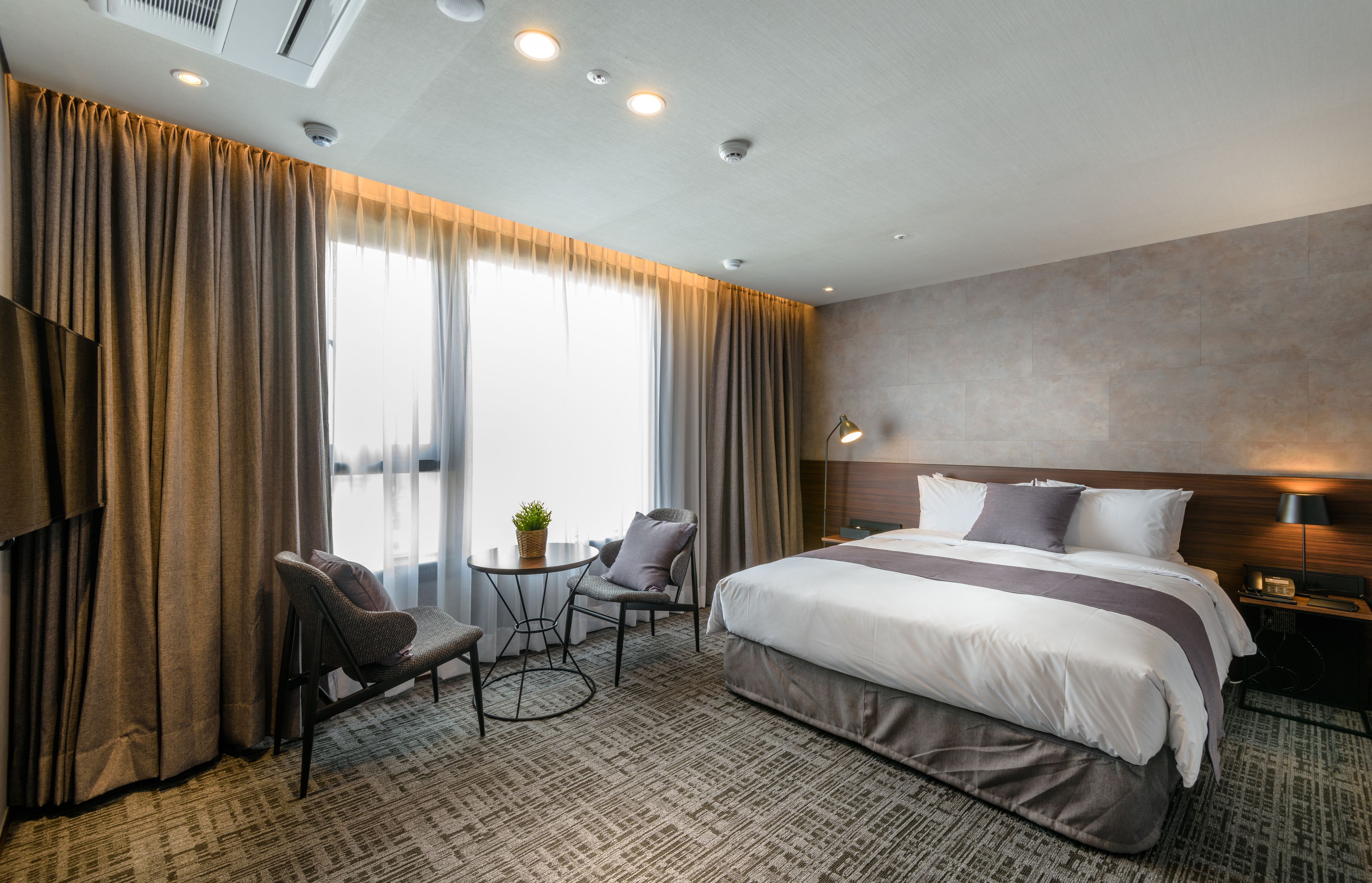 A hotel room with a large bed, a desk with a chair, a small table with a plant, and a large window with curtains.