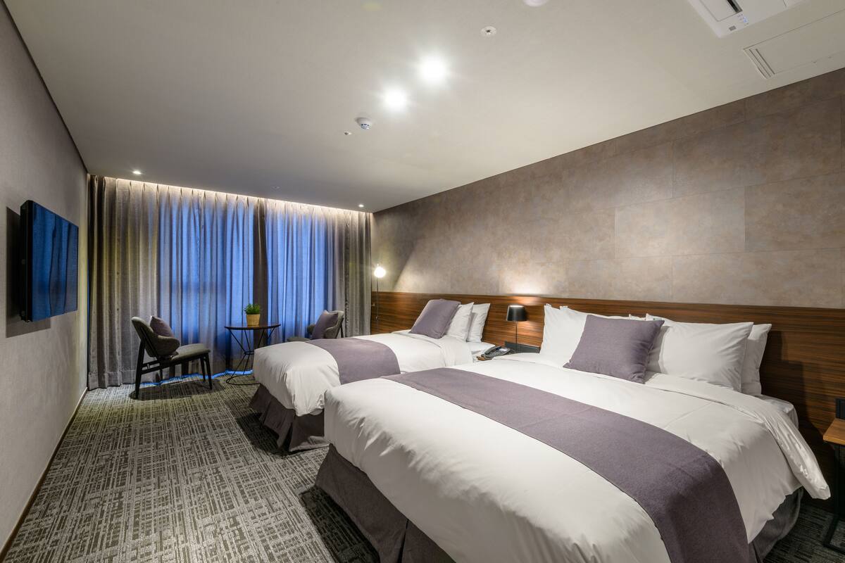 Une chambre d’hôtel avec deux lits, un bureau, une chaise, une petite table et une grande fenêtre avec des rideaux.
