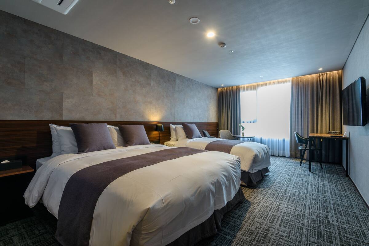 Une chambre d’hôtel avec deux lits, un bureau, une chaise, un téléviseur à écran plat et une grande fenêtre avec des rideaux.