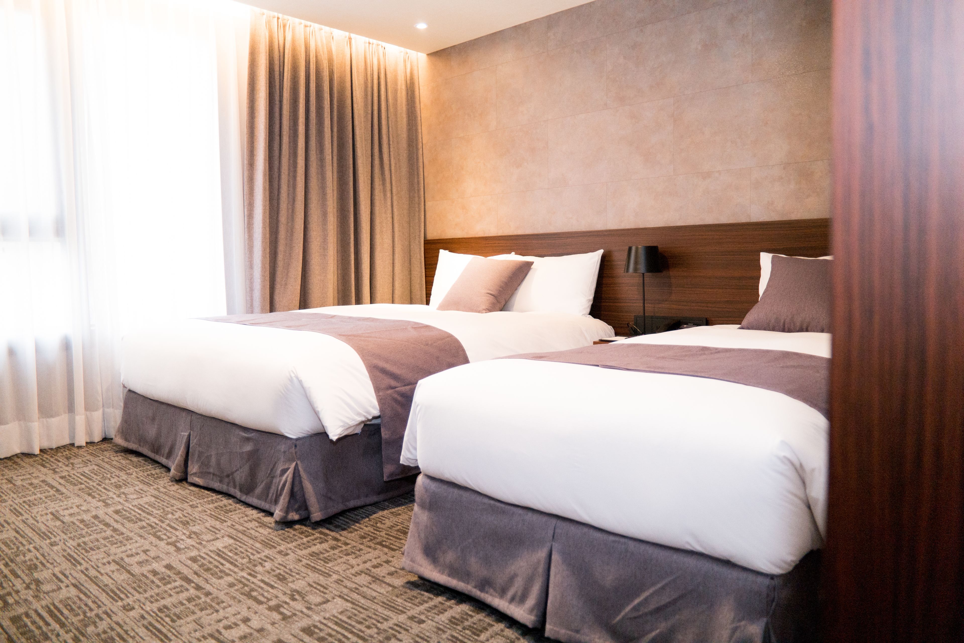 A hotel room with two beds, a large window with curtains, a carpeted floor, and a wooden headboard.