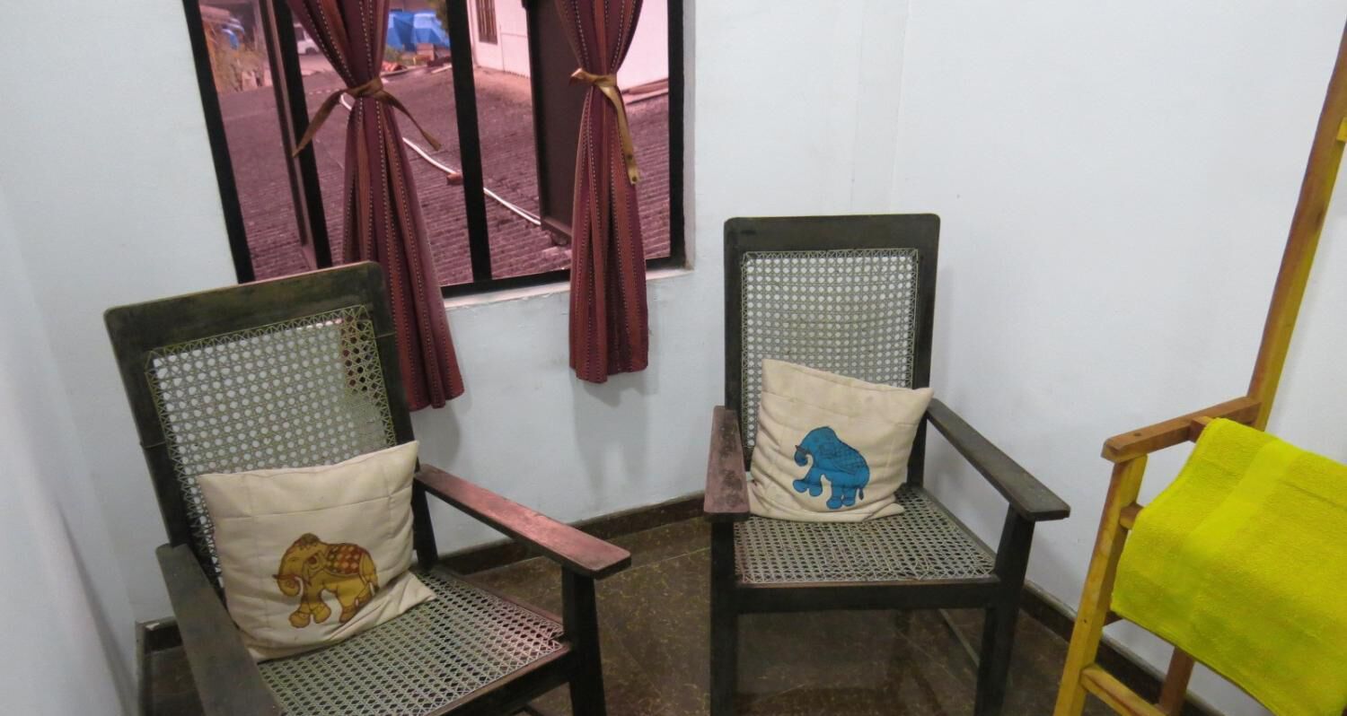 Two chairs with mesh backs and cushions, one with a yellow elephant and the other with a blue elephant, against a white wall with a window and curtains.