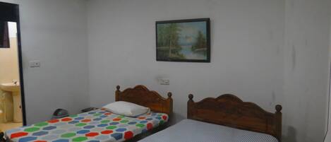 Una habitación pequeña y sencilla con dos camas individuales, un cabecero de madera, un cuadro en la pared y un lavamanos en una esquina.