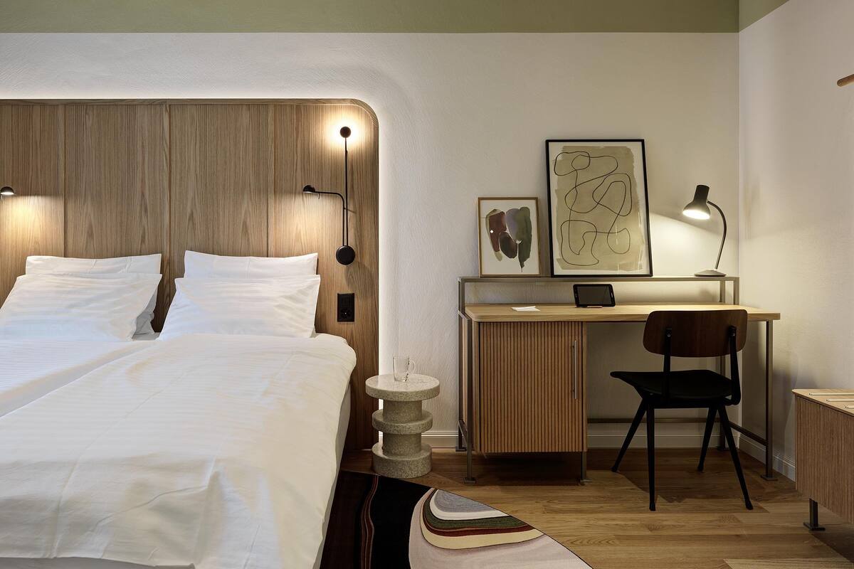 A neatly arranged bedroom with a wooden headboard, a bedside table, a desk with a chair, and a bed with white bedding.