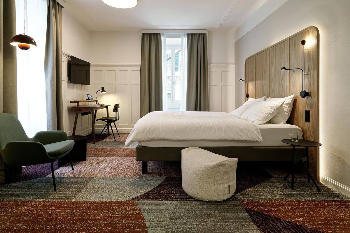 A modern hotel room with a large bed, a desk, a chair, and a small table with a lamp.