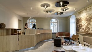 A modern reception area with a curved wooden desk, a receptionist, and a large window. - Hotel St Josef (Zürich)