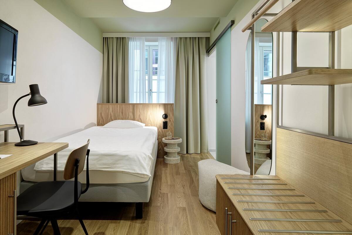 A modern hotel room with a wooden bed, a desk with a chair, a small round table, and a mirror.
