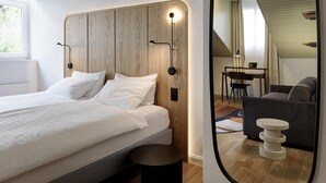 A modern bedroom with a wooden headboard, a round mirror, a bed with white linens, a small round table, and a grey sofa. - Hotel St Josef (Zürich)