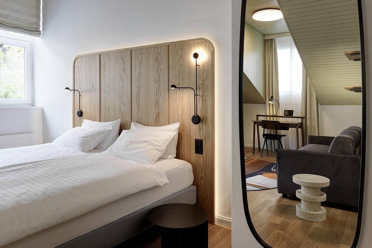 A modern bedroom with a wooden headboard, a round mirror, a bed with white linens, a small round table, and a grey sofa.