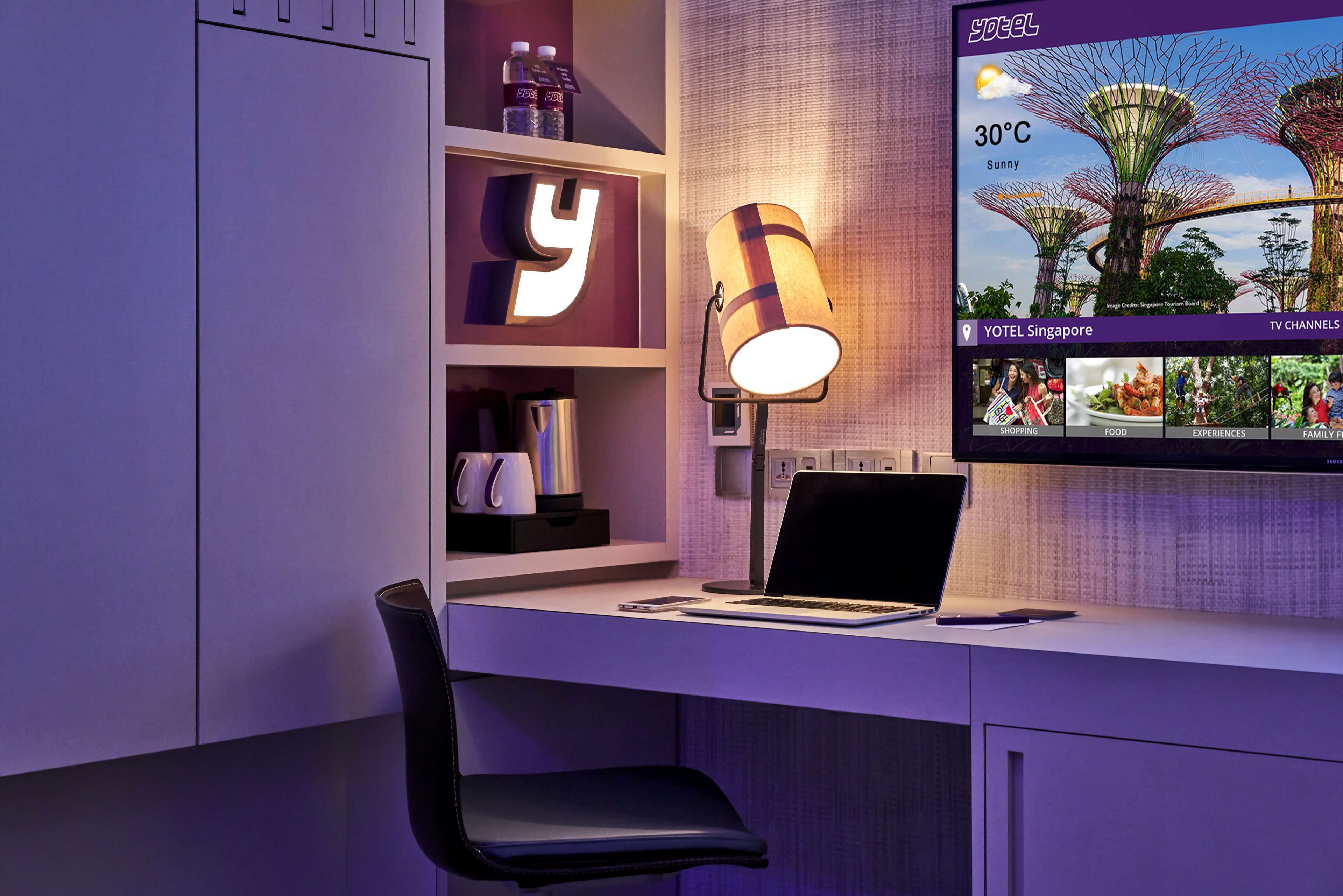 Une chambre d’hôtel moderne équipée d’un bureau, d’un ordinateur portable, d’une machine à café et d’un téléviseur diffusant des images d’un paysage tropical.
