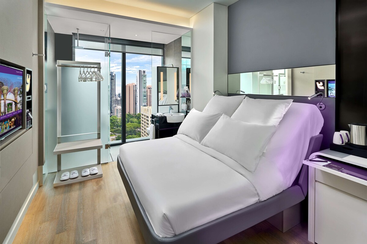 A modern hotel room with a large bed, a vanity with a mirror, a desk with a chair, and a view of the city through large windows.