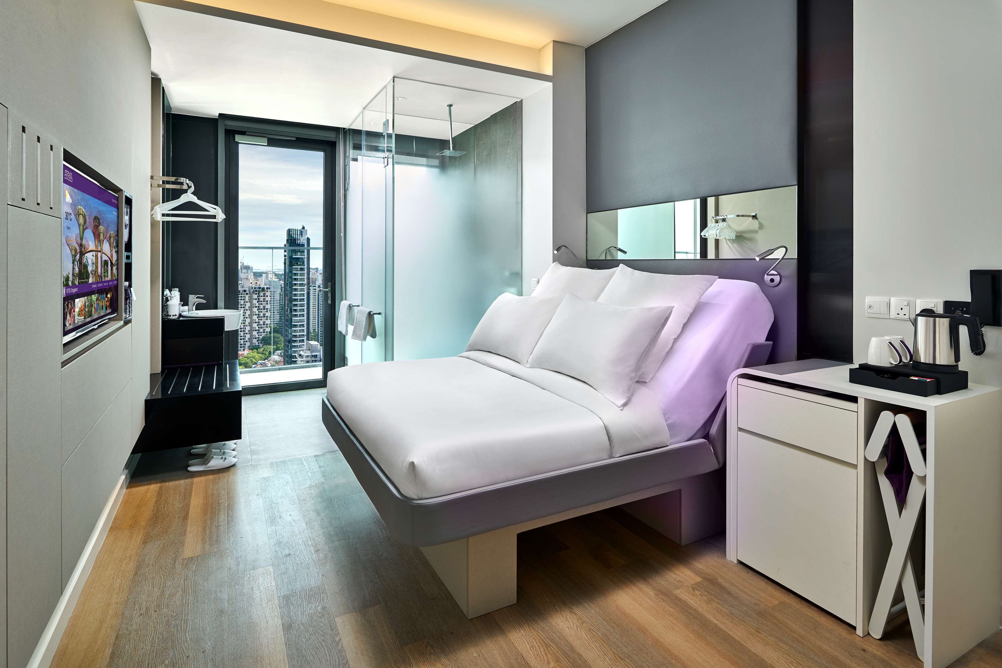 A modern hotel room with a bed, a desk, a mirror, and a view of the city.