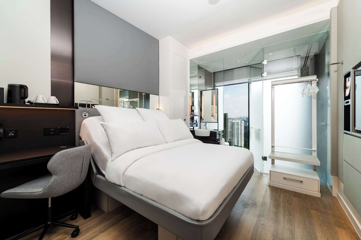 A modern hotel room with a bed, desk, chair, and a view of the city through a glass door.