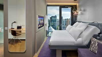 A modern hotel room with a large bed, a desk with a chair, a large mirror, and a city view.