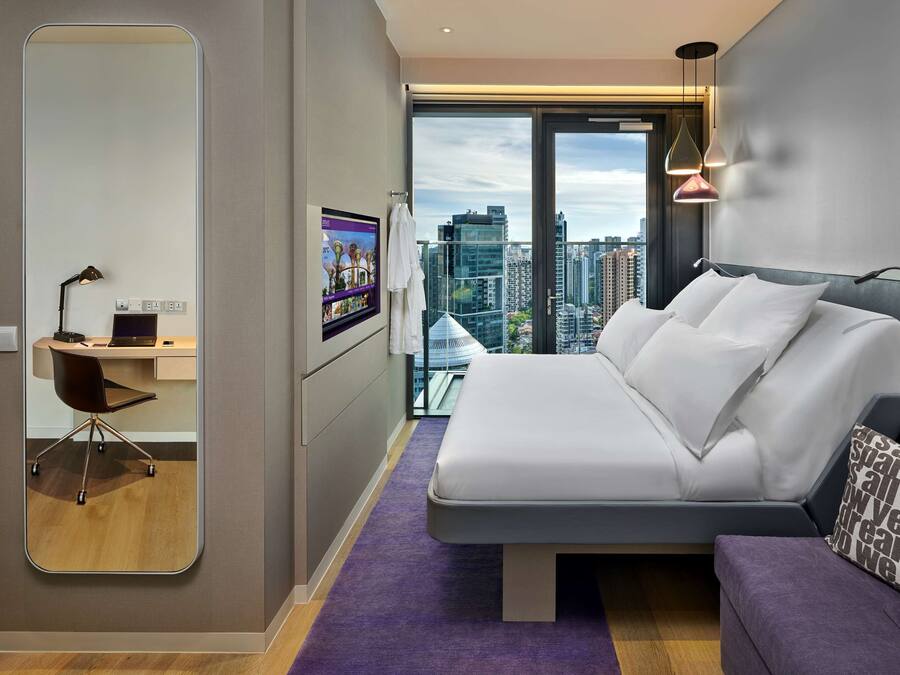 A modern hotel room with a large bed, a desk with a chair, a large mirror, and a city view.