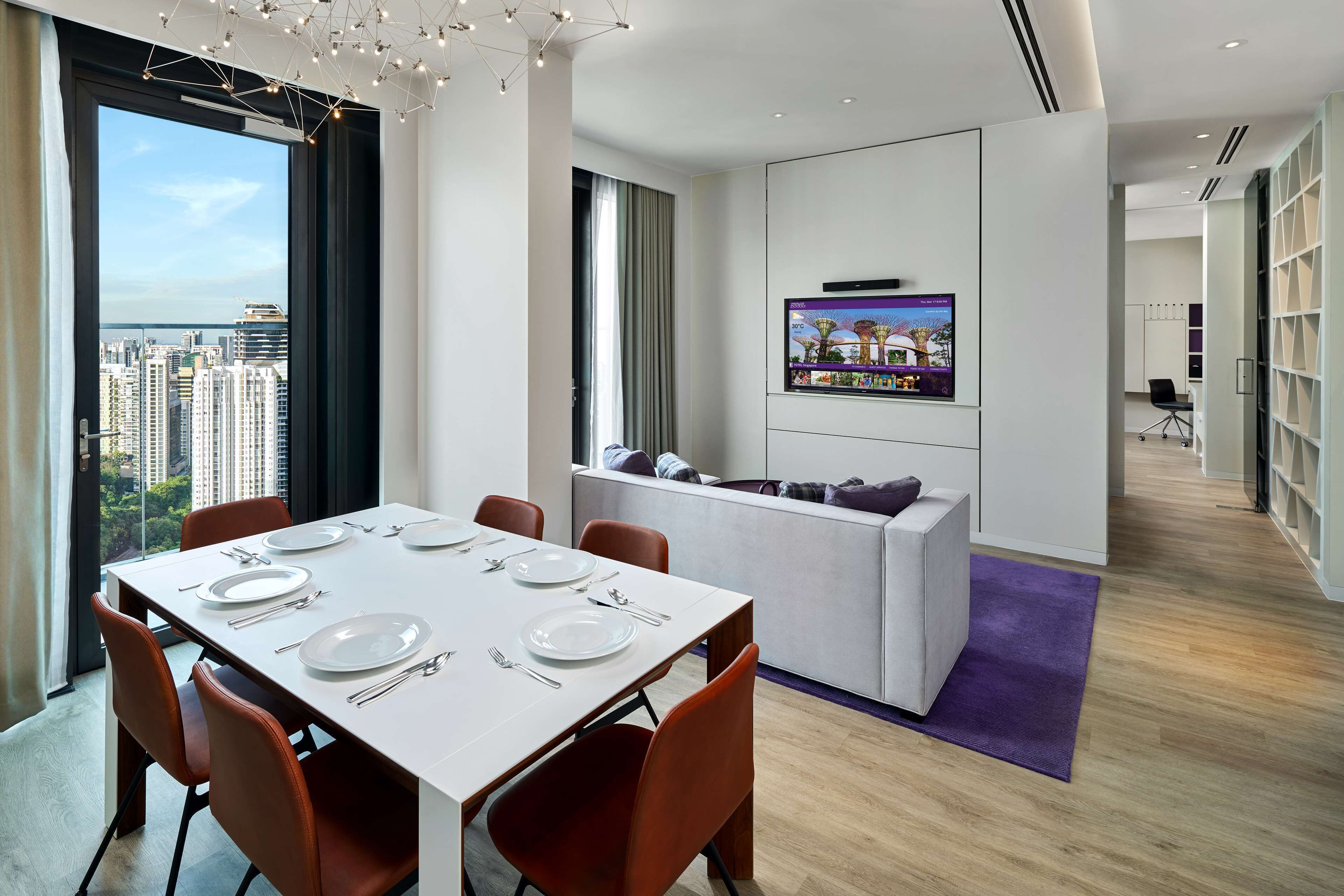 A modern hotel room with a dining area, a seating area, a TV mounted on the wall, and a view of the cityscape.