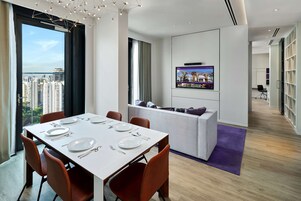 A modern hotel room with a dining area, a seating area, a TV mounted on the wall, and a view of the cityscape.