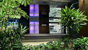 A modern building entrance with a glass facade, a sign reading 'HOTEL', and lush greenery in the foreground.