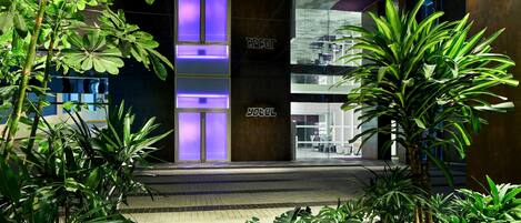 A modern building entrance with a glass facade, a sign reading 'HOTEL', and lush greenery in the foreground.