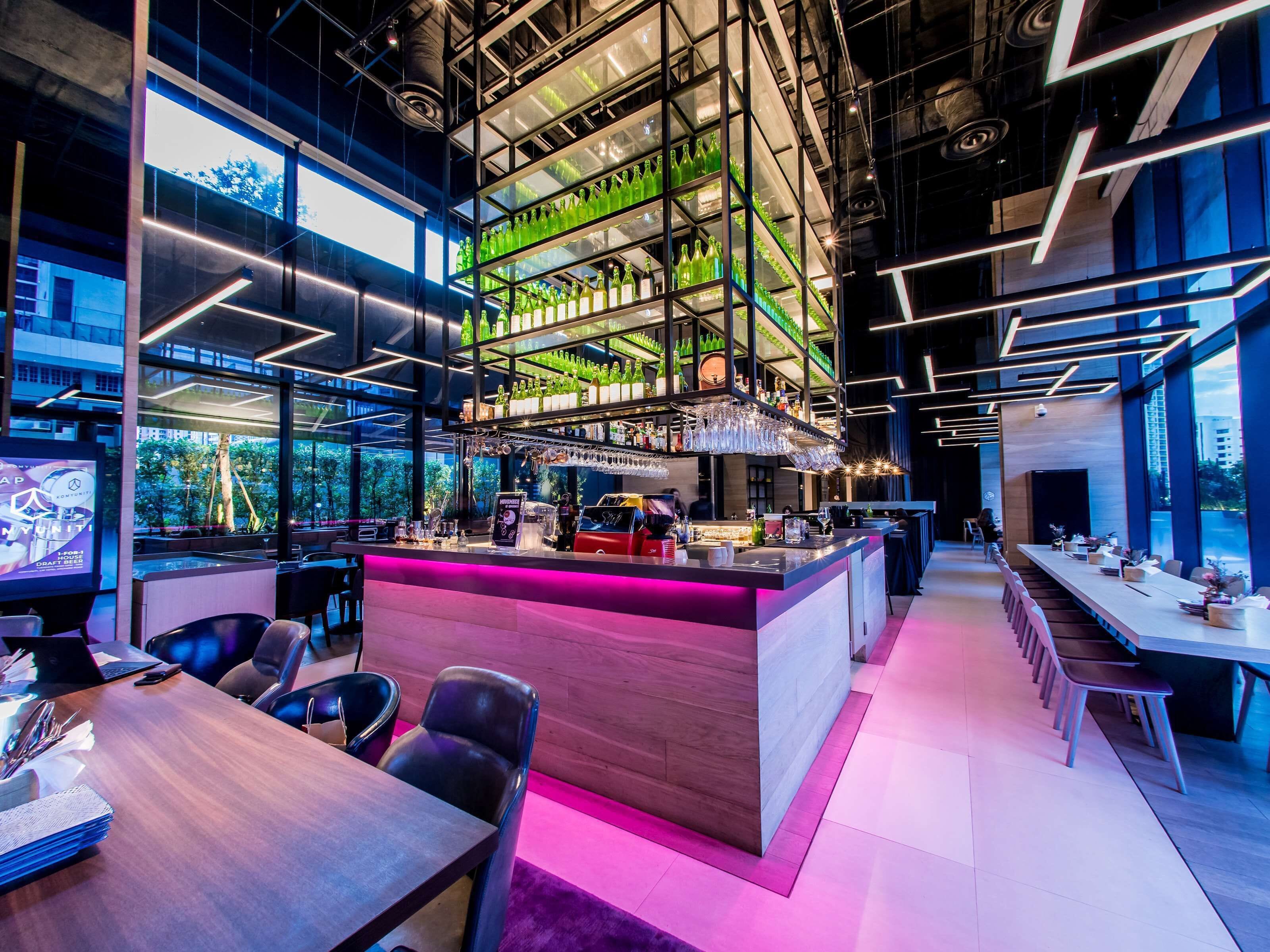 A modern bar area with a high ceiling, shelves of green bottles, and a long counter with bar stools.