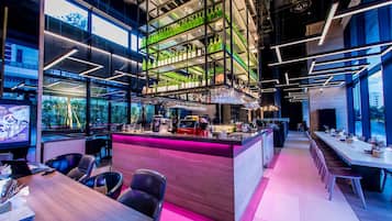 A modern bar area with a high ceiling, shelves of green bottles, and a long counter with bar stools.