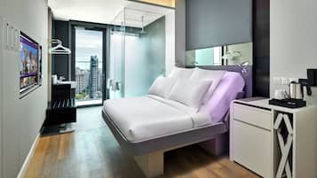 A modern hotel room with a bed, a desk, a mirror, and a view of the cityscape.