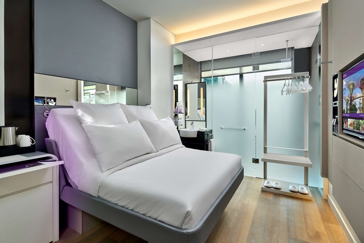 A modern hotel room with a large bed, a bedside table, a mirror, a wardrobe, and a shoe rack.