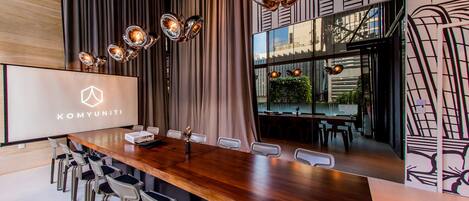 A conference room with a long wooden table, modern chairs, and a large screen displaying the logo "KOMYUNITI".