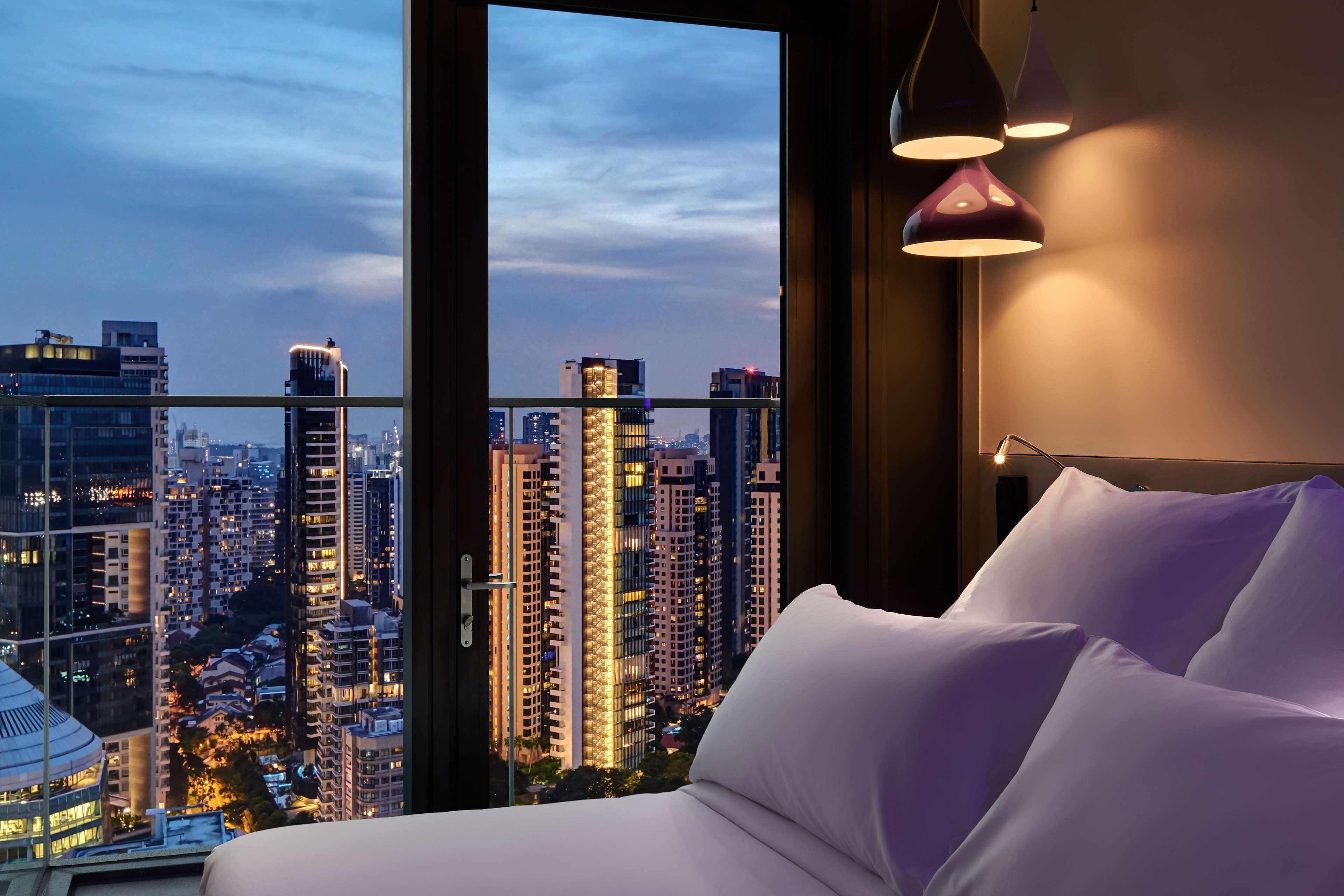 A hotel room with a large window offering a cityscape view, a bed with white bedding, and a modern pendant light fixture.