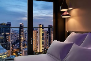 Habitación de hotel con un ventanal grande que ofrece vistas panorámicas de la ciudad, una cama con ropa blanca y una lámpara colgante de diseño moderno.