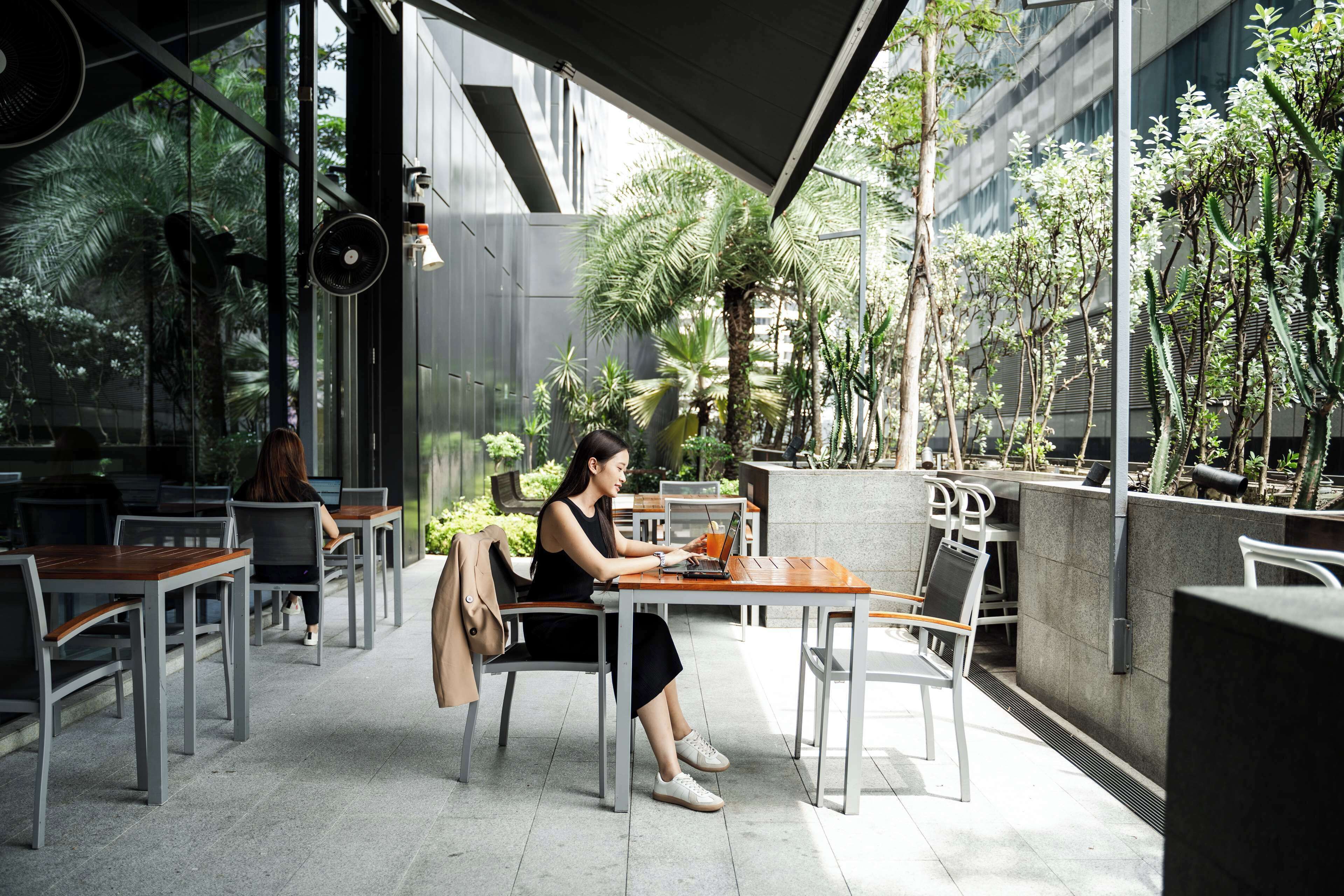 Een vrouw zittend aan een tafel met een laptop in een modern café met buitenruimte, glazen wanden en planten.