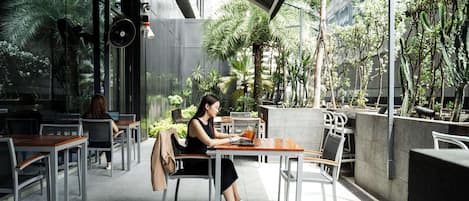 Una mujer sentada a una mesa con una computadora portátil en un moderno café al aire libre con paredes de vidrio y plantas.