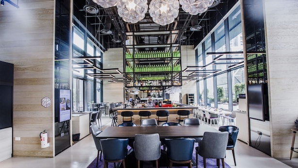 A modern dining area with a long wooden table, multiple chairs, and a bar in the background. - YOTEL Singapore Orchard Road (Singapore)