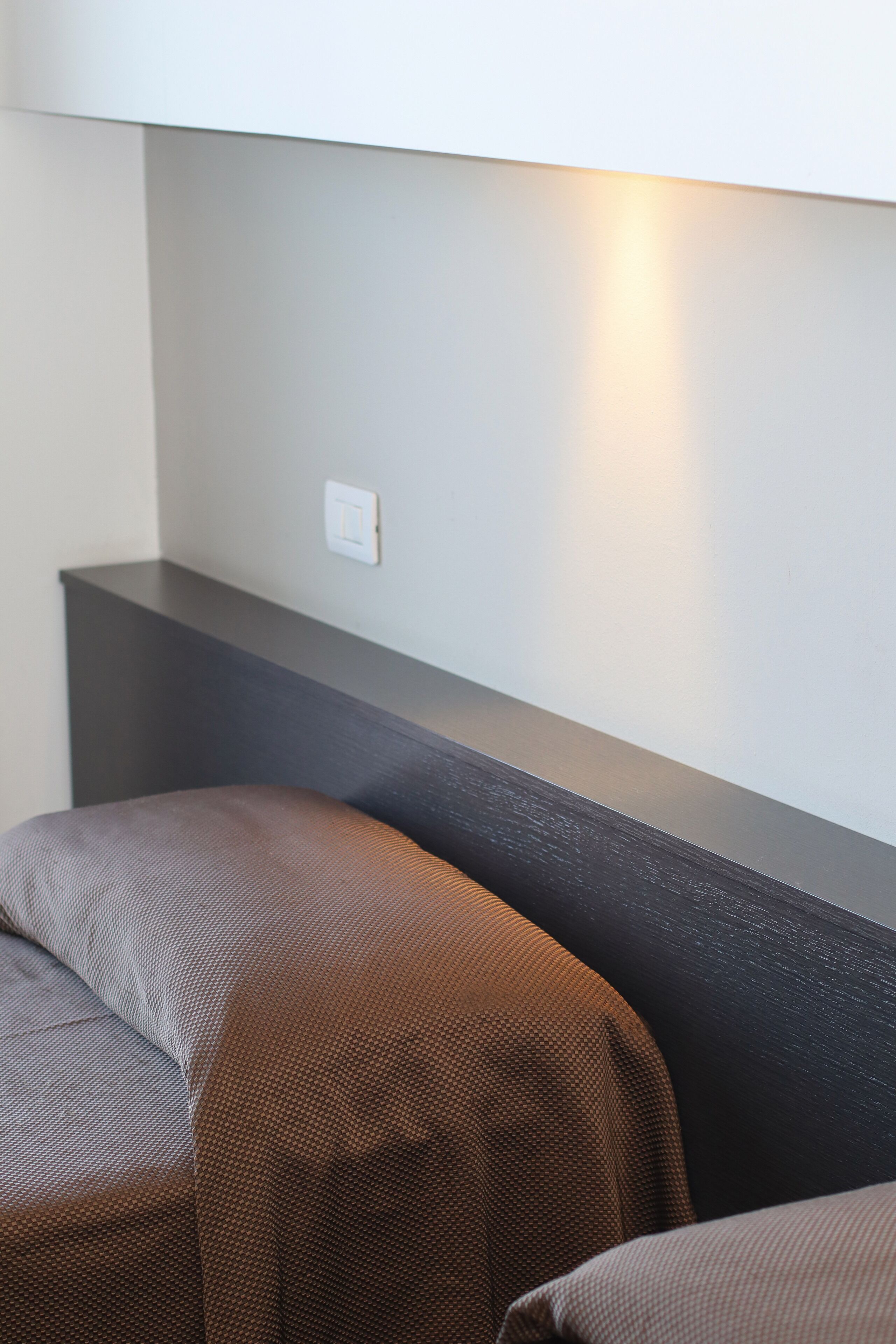 Habitación de hotel con cabecera oscura, un interruptor de luz en la pared y una cama parcialmente visible con colcha estampada.