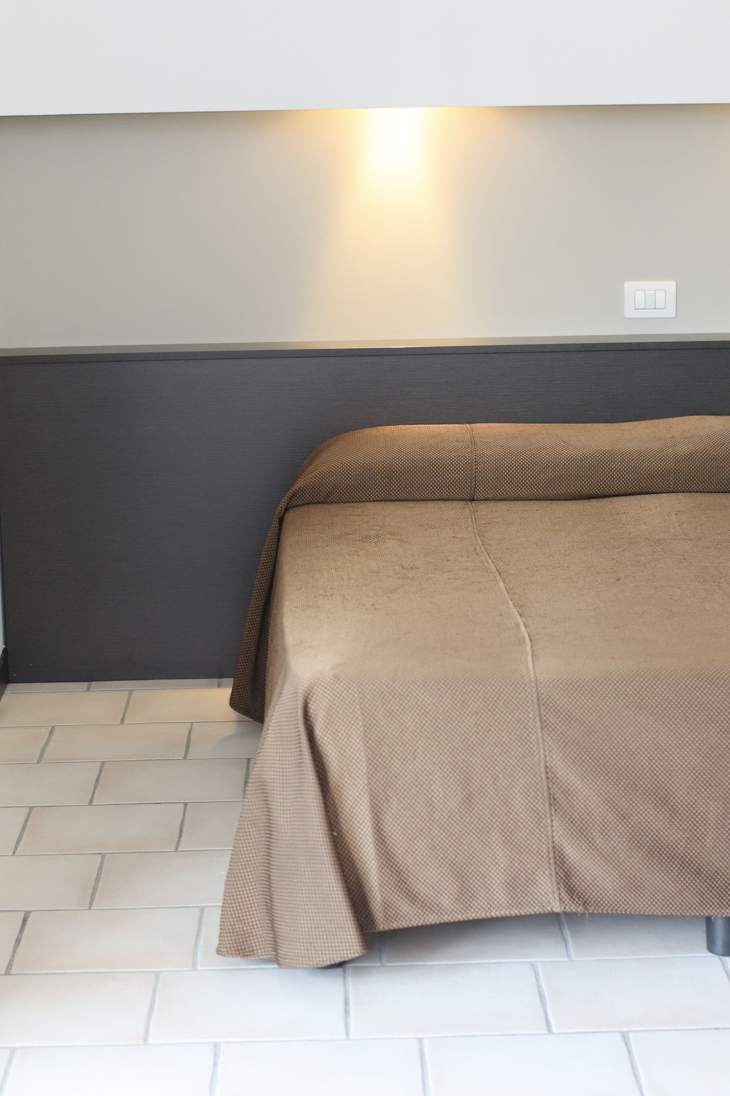 A bed with a brown cover in a room with a light-colored floor and a dark wall.