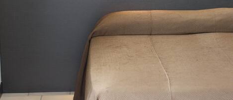 Un lit recouvert d’une couverture marron, dans une pièce dont le sol est de couleur claire et un des murs est foncé.